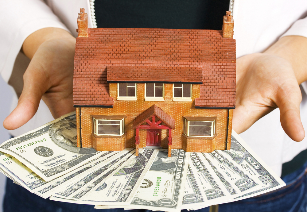 A person holding a miniature house and some dollar bills for making payments